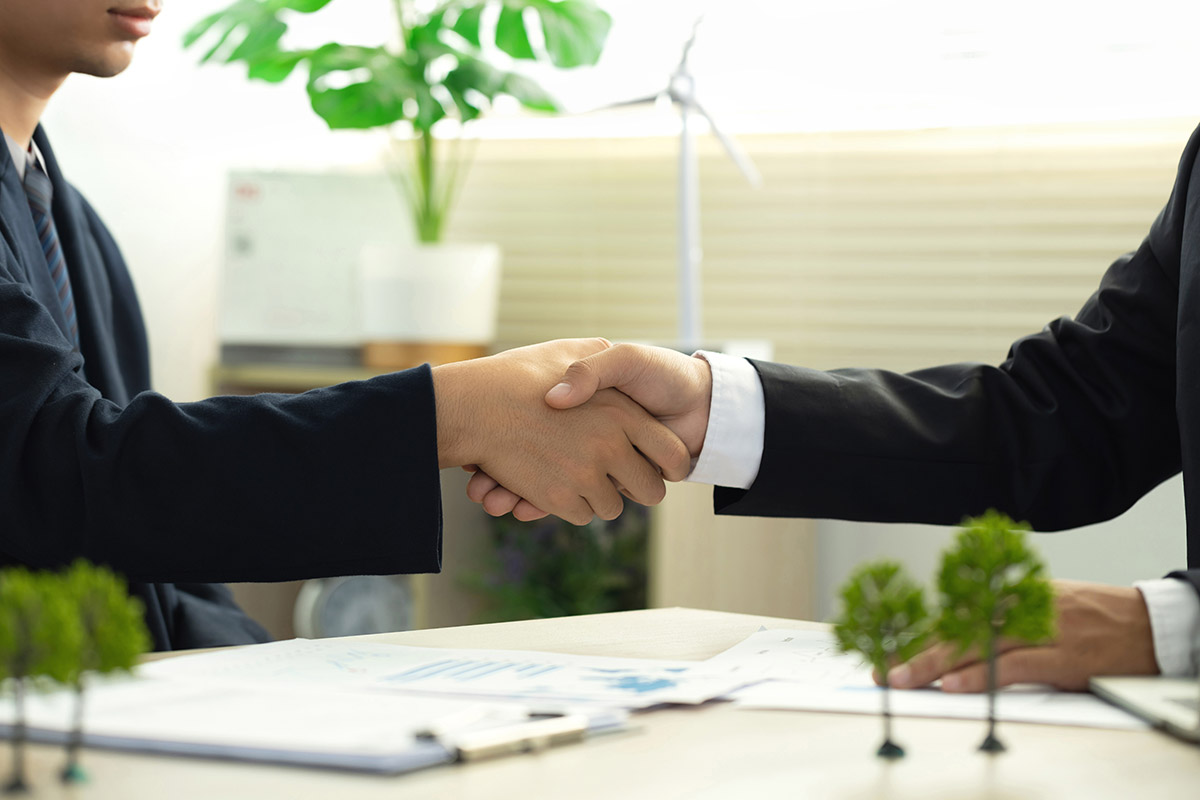 Two business professionals shake hands over a table with ESG (Environmental, Social, and Governance) documents and green energy models.